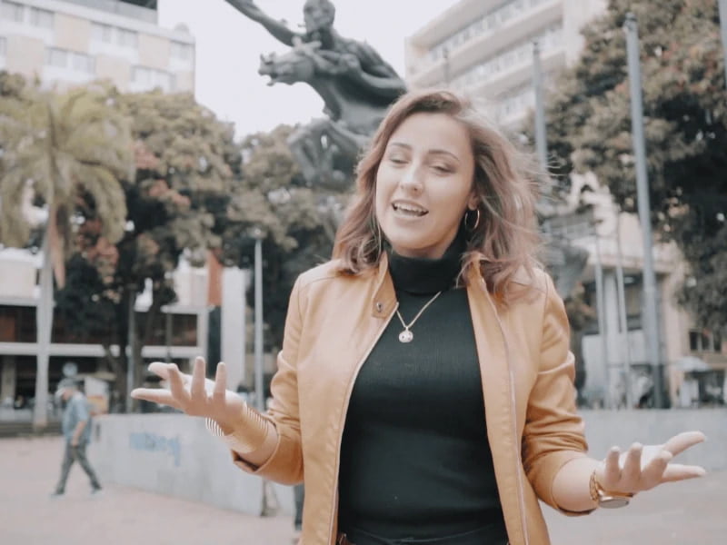 Erika Paola Camargo Monroy is captured in a lively outdoor setting, gesticulating animatedly with a joyful expression on her face. She is dressed in a stylish mustard-yellow jacket over a black turtleneck. The backdrop features an urban square with a large, dramatic statue and several high poles. Trees and city buildings can be seen in the soft focus background, enhancing the lively atmosphere of the public space where she stands. This dynamic scene is brought to you by the Architectural Phoenix Team, showcasing not just our designs but the vibrant urban life they foster.