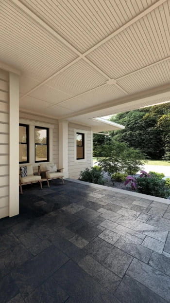 A modern outdoor patio area with dark stone flooring, light-colored wooden walls, and a white wooden ceiling. Two beige cushioned chairs are placed against the wall on the left side, and the view outside reveals a lush green garden with various shrubs and trees.