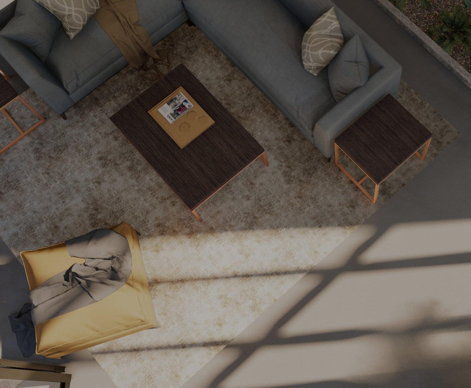 Overhead view of a contemporary living room featuring a dark gray sectional sofa adorned with patterned throw pillows. The central area includes a large beige area rug and a dark wooden coffee table, accompanied by two matching side stools. A golden pouf adds a splash of color. Natural light casts geometric shadows across the room, creating a cozy and inviting atmosphere