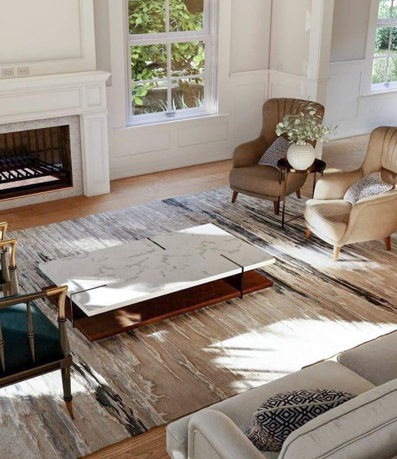 Spacious living room featuring a traditional fireplace with a white mantel, surrounded by large windows offering views of greenery. The room is furnished with two beige upholstered armchairs with black and white patterned pillows, a modern white marble coffee table with a wooden base, and a large, abstract-patterned area rug in shades of beige and gray. Sunlight filters through the windows, creating a warm and inviting atmosphere.